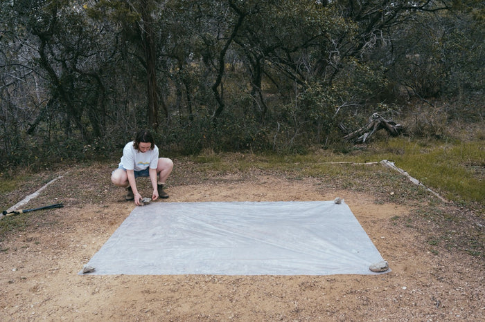 Gossamer Gear Polycro Ground Cloth laid flat on dirt, hiker staking a corner