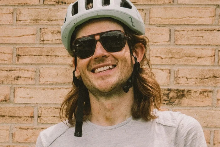 Ombraz Leggero armless sunglasses worn by a smiling cyclist in a helmet by a brick wall
