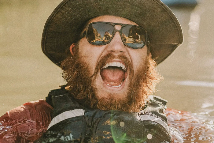 Ombraz Leggero armless sunglasses worn by a laughing man in a hat, chest-deep in water