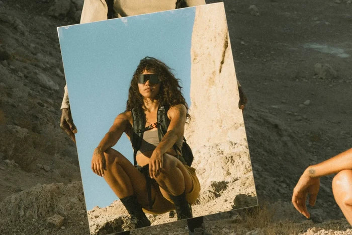 Ombraz Refugio armless sunglasses reflected in a mirror held by a crouching woman
