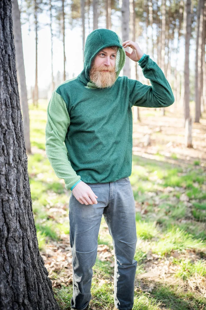 Sambob Alpha 90 Hoodie in Arbor/Fern – worn hood up by a hiker beside a pine trunk in a forest