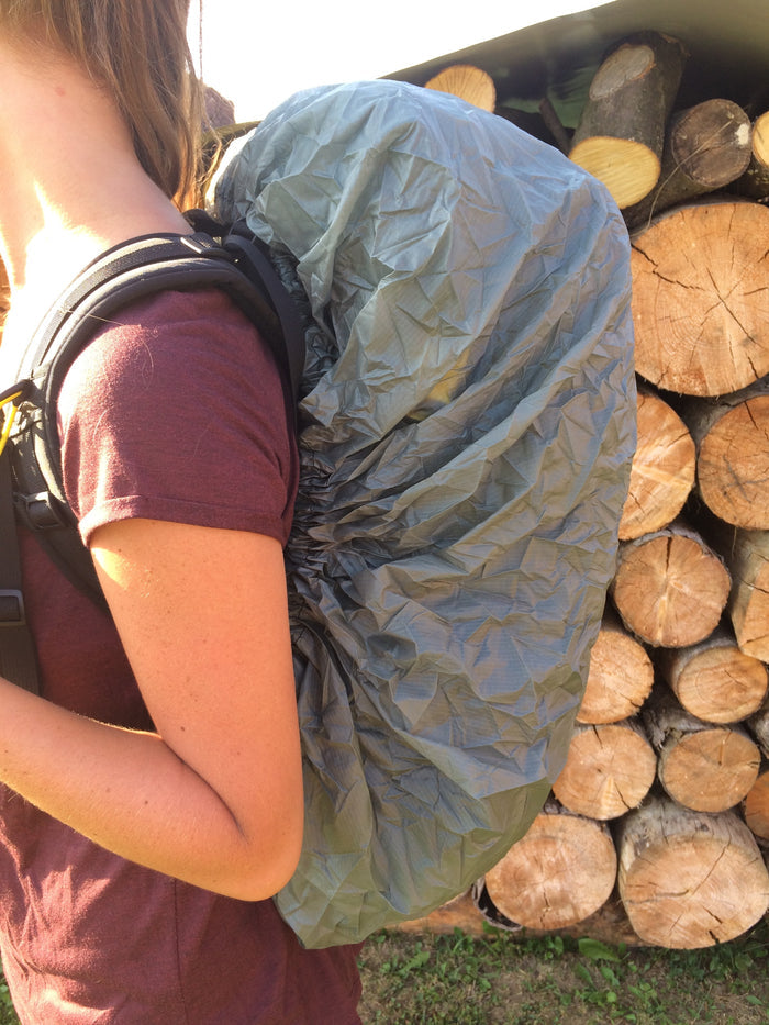 ULA Pack Cover in grey fitted over a pack worn by a hiker, woodpile behind